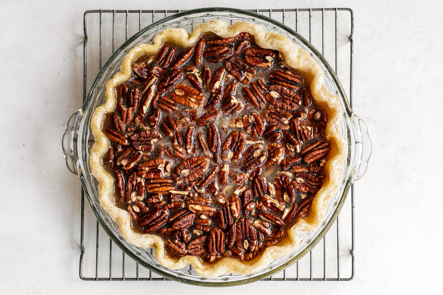 overhead shot of a whole, unsliced pecan pie in a glass pie pan on top of a cooling rack.